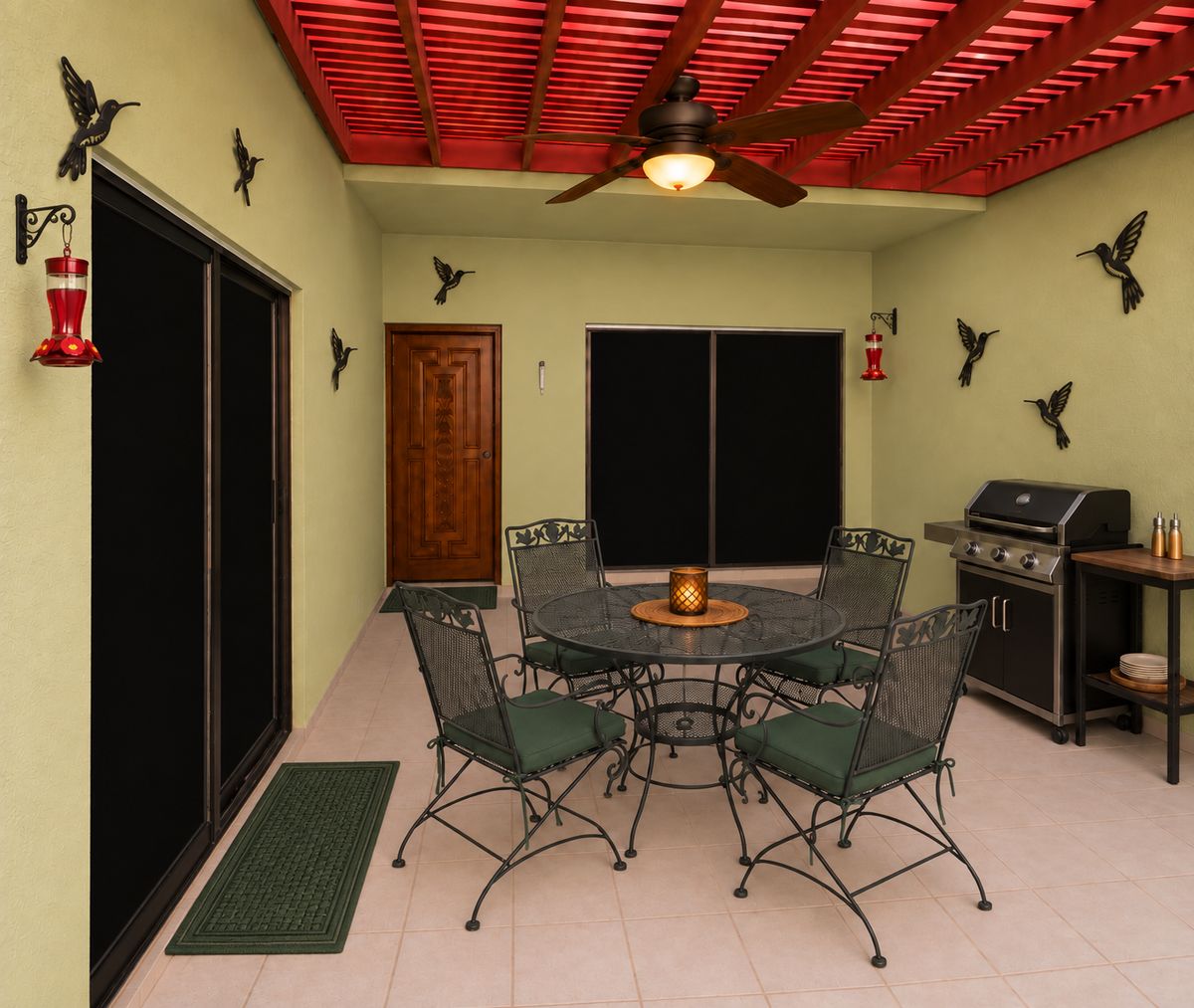 The covered patio with a red pergola ceiling, wrought-iron dining set, a gas grill and hummingbird wall art