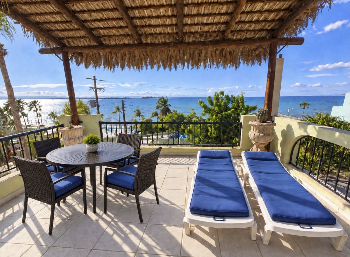 The Ocean Suite balcony under a palapa roof: a dining table, two blue loungers and a wide view of the Bay of La Paz